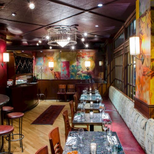 A cozy bar with a long dining table setup, wine bottles on a shelf, warm lamps, and artwork along the walls, ready for guests tonight.