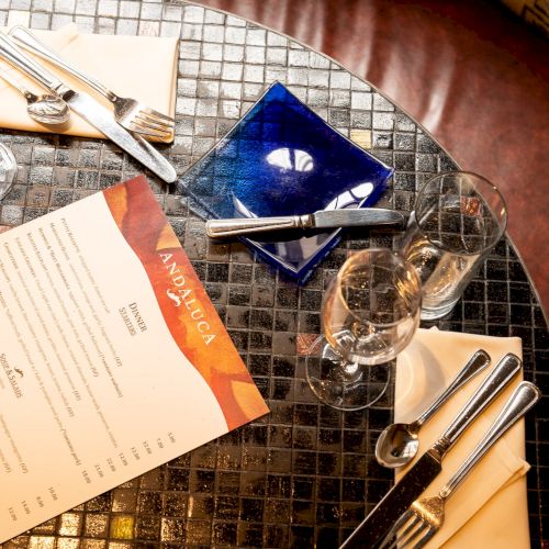 A round table set for two with wine glasses, a menu, cutlery, napkins, a blue ashtray, and a glossy tiled surface.