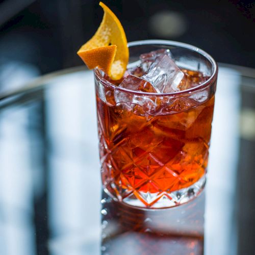 A glass of dark amber cocktail with ice, garnished with an orange twist, on a reflective surface (top it at 140 characters, always ending the sentence).