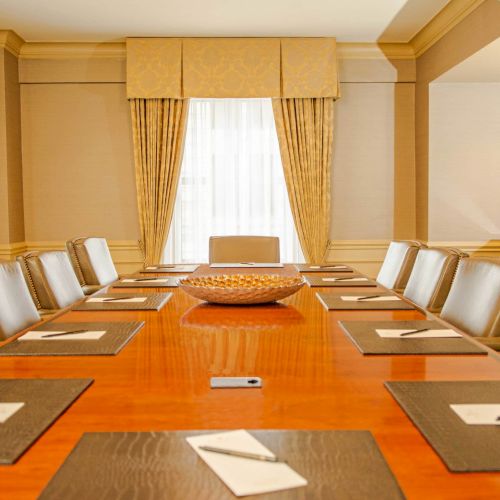 A long polished wooden conference table with leather chairs, place settings, notepads, a central bowl, and elegant beige‑golden decor.