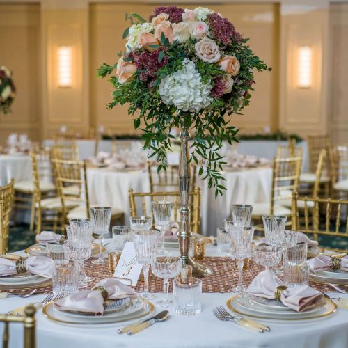 A lavish wedding reception setup with a tall floral centerpiece, elegant round tables, white linens, gold accents, and warm ambient lighting.