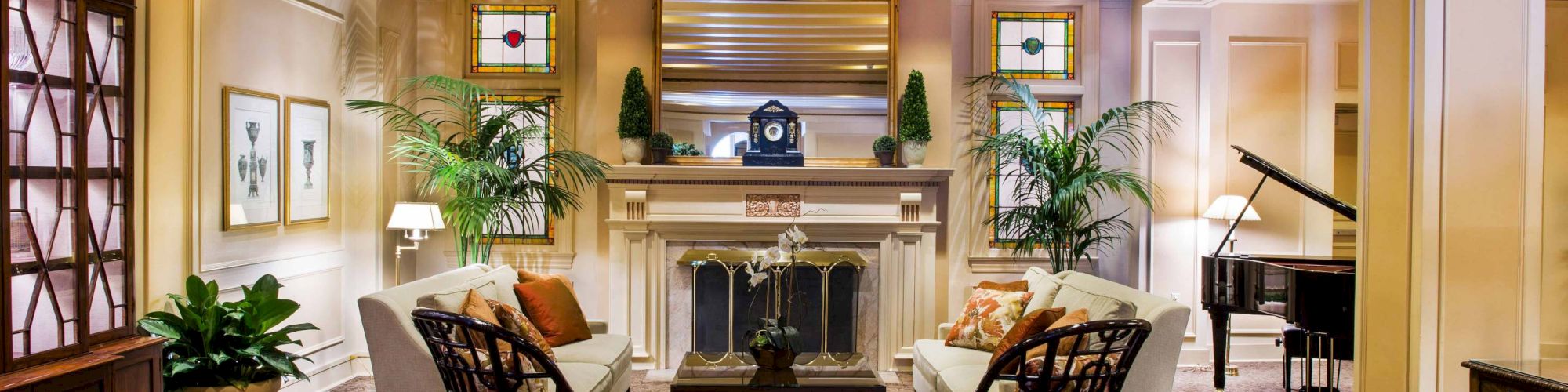 A cozy hotel lobby with a central fireplace, two upholstered sofas facing each other, wicker chairs, potted plants, soft lighting, and a grand piano in the back right, all under a warm, inviting glow.
