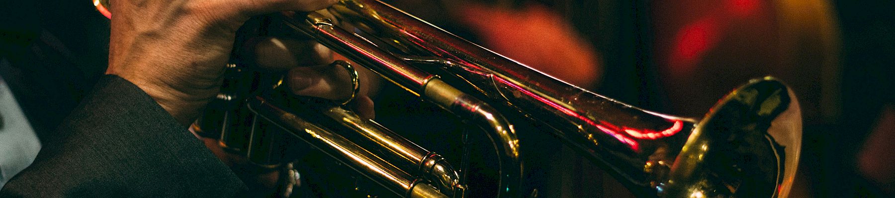 A close-up of a musician's hands playing a brass trumpet, dim stage lighting in the background, capturing a live jazz vibe.