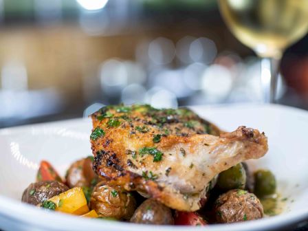 A plated roasted chicken leg on a bed of colorful roasted vegetables, garnished with herbs and greens, with a glass of white wine in the background.