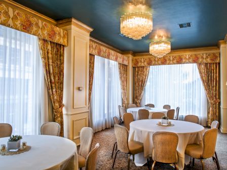 A bright, elegant dining room with round tables, beige chairs, large windows with draped curtains, and chandeliers overhead.