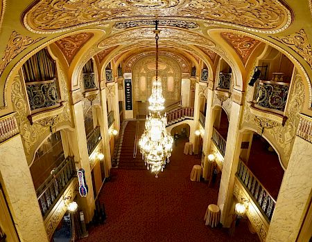 A grand, ornate theater lobby with gilded arches, balconies, and a large chandelier, viewed from above.