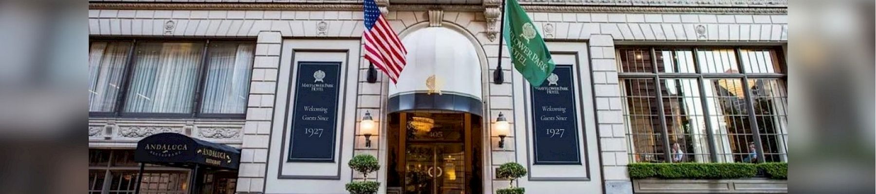 A grand hotel entrance with American and green flags, stone facade, and steps leading to a revolving door. (140 chars max, ending with a period)