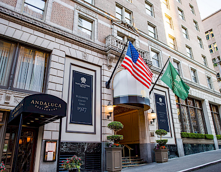 A hotel facade with U.S. and green state flags, showing signs for Mayflower Park Hotel and Andalucca Restaurant.