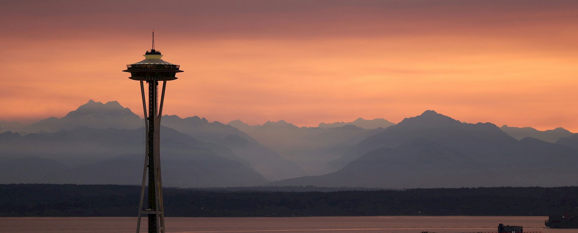 Seattle skyline at dusk with the Space Needle silhouetted against a fiery orange sky over the water, distant mountains, and city lights.