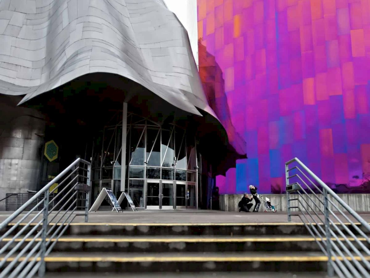 A futuristic building with curving gray and vibrant purple-orange walls, metal steps leading to glass doors, and a few people nearby.