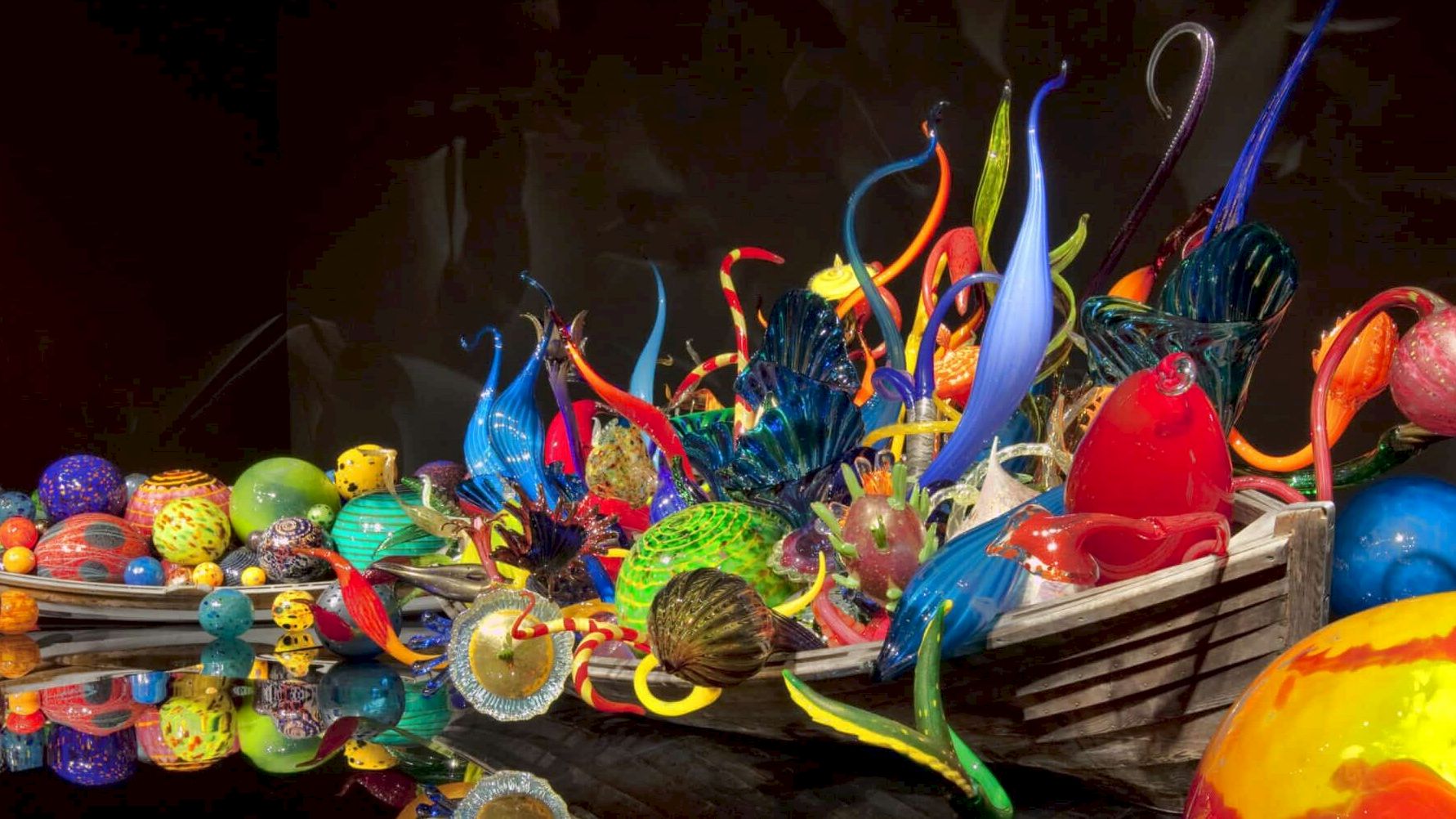 A vibrant glass art sculpture boat filled with colorful glass shapes and swirls, reflected in dark water&mdash;bright chaos on a calm surface.