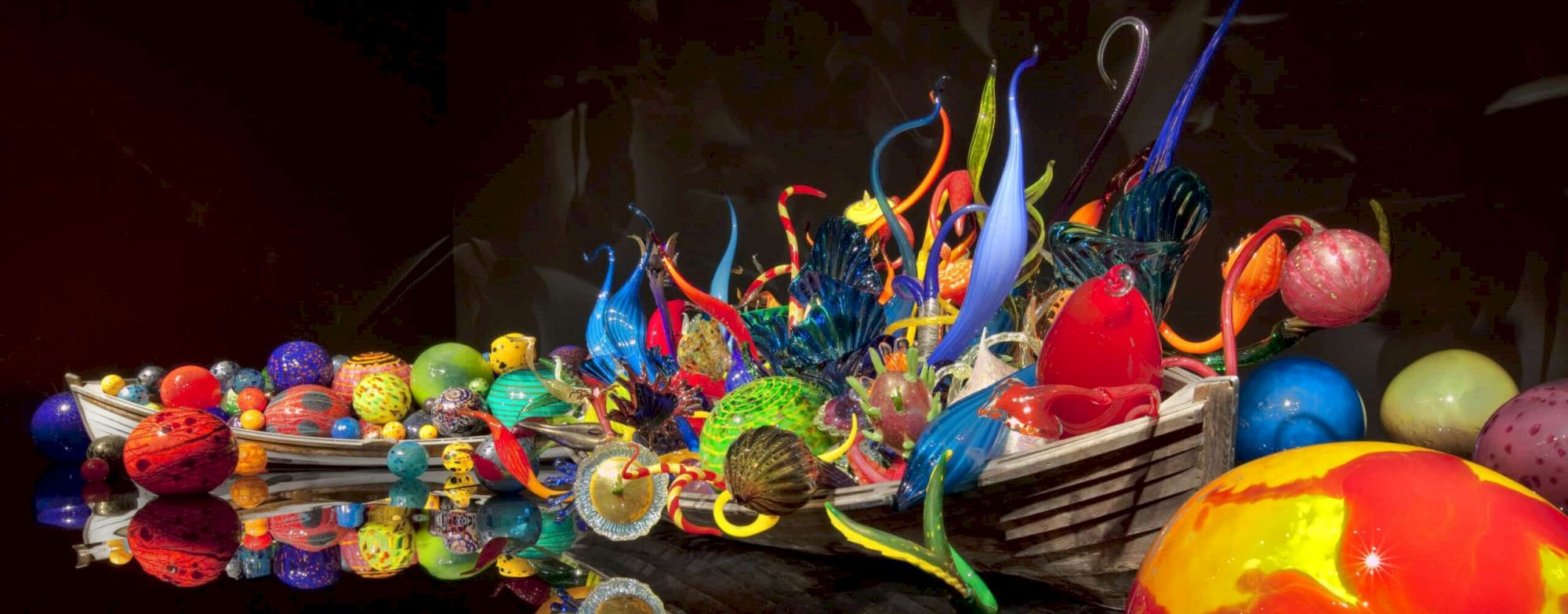 A vibrant glass art sculpture boat filled with colorful glass shapes and swirls, reflected in dark water&mdash;bright chaos on a calm surface.