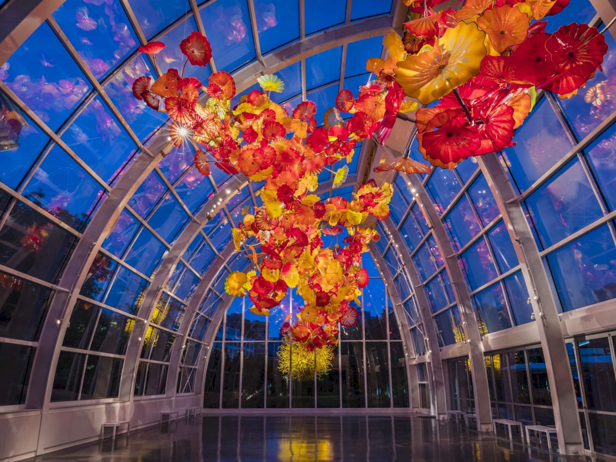 A glass greenhouse with a curved, arched ceiling, displaying vibrant hanging autumn leaves in red, orange, and yellow.