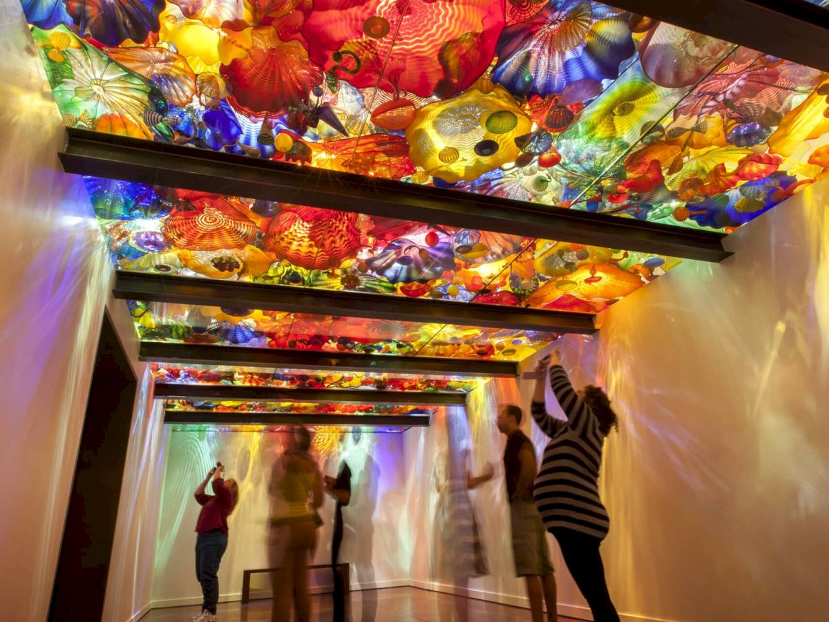 People walking through a long, colorful ceiling art tunnel of glowing, flower- and pattern-covered panels, with reflections on the glossy floor.