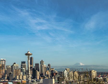 A city skyline with tall buildings and a prominent tower, under a bright blue sky with a distant mountain peak on the horizon.