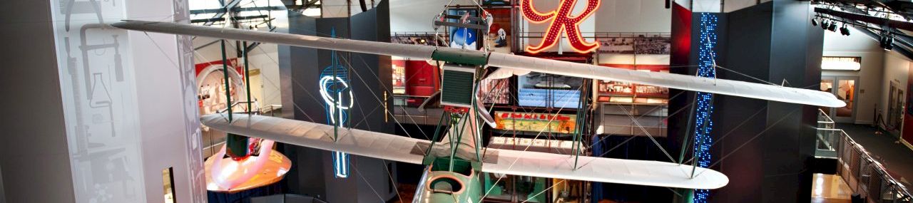 A large indoor display space with model airplanes hanging from the ceiling, a red &ldquo;R&rdquo; sign, and toy vehicles scattered on a polished wooden floor.