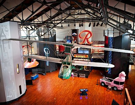 A large indoor display space with model airplanes hanging from the ceiling, a red &ldquo;R&rdquo; sign, and toy vehicles scattered on a polished wooden floor.