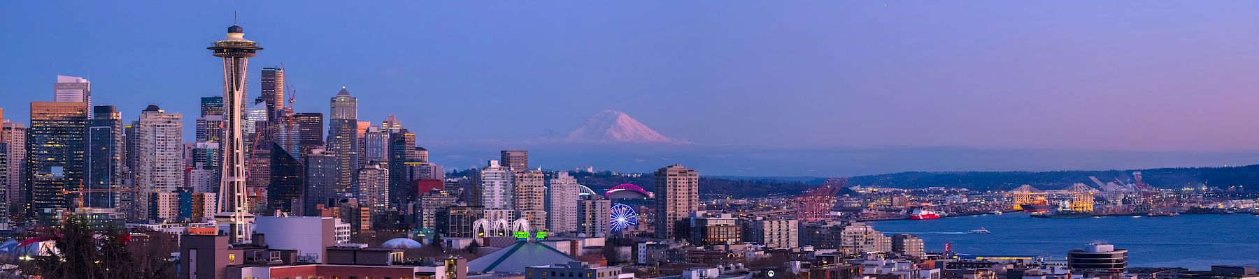 A city skyline with tall buildings and Space Needle-like tower overlooking a waterfront at dusk, with a distant mountain in the background.