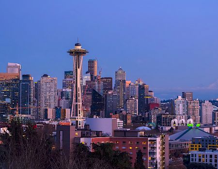 A city skyline with tall buildings and Space Needle-like tower overlooking a waterfront at dusk, with a distant mountain in the background.