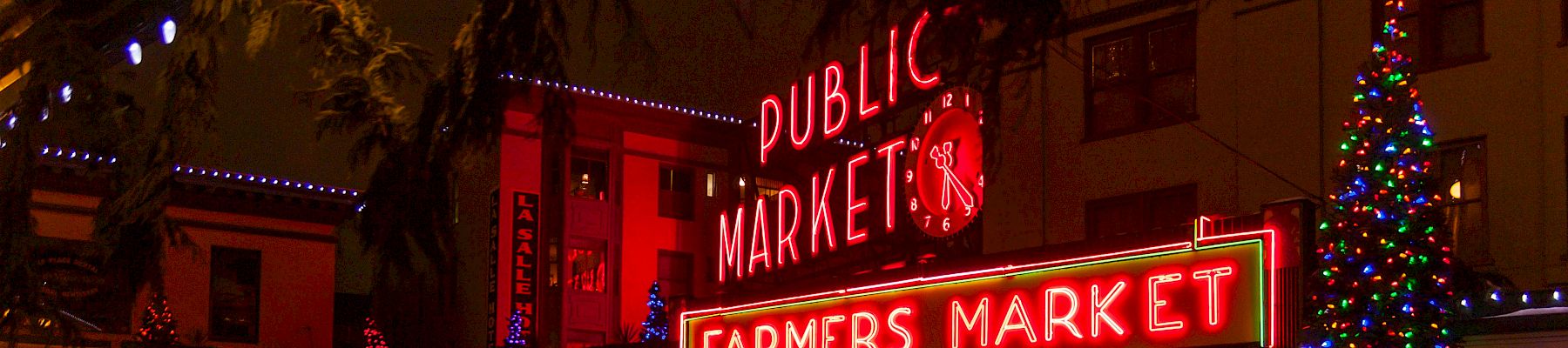 A festive outdoor market scene at night with bright red &ldquo;Public Market&rdquo; and &ldquo;Farmers Market&rdquo; neon signs, tree lights, and a glowing plaza.