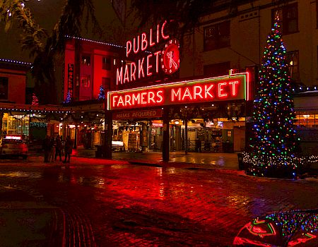A festive outdoor market scene at night with bright red &ldquo;Public Market&rdquo; and &ldquo;Farmers Market&rdquo; neon signs, tree lights, and a glowing plaza.