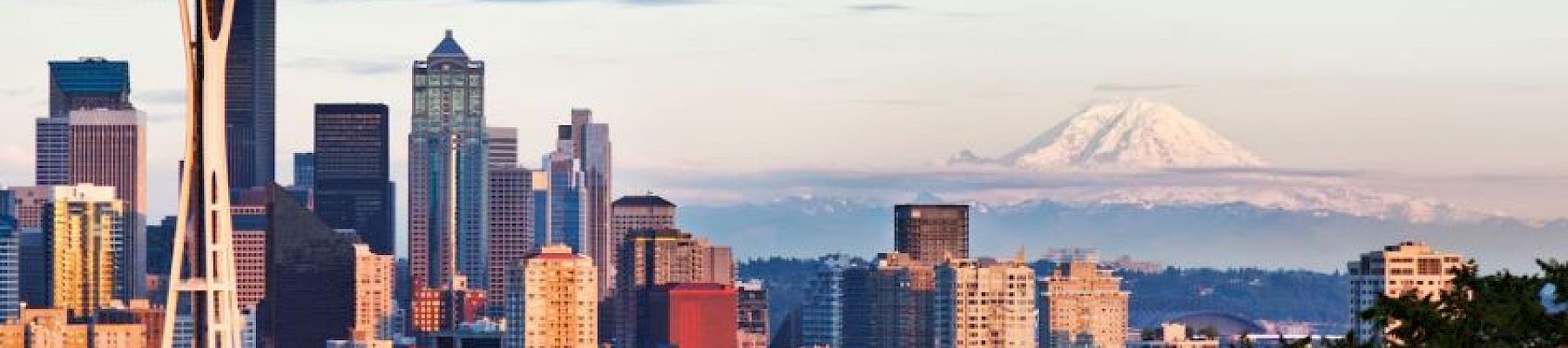 Seattle skyline with Space Needle in foreground and mountains (possibly Mount Rainier) in the distance at dusk.