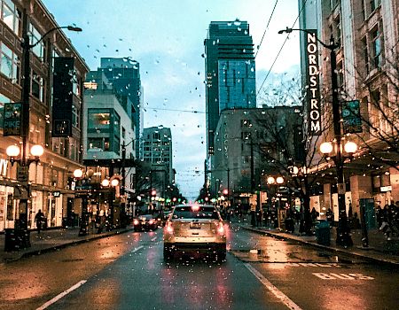 A city street at dusk with wet pavement, cars in traffic, storefronts lit up, and string lights overhead.