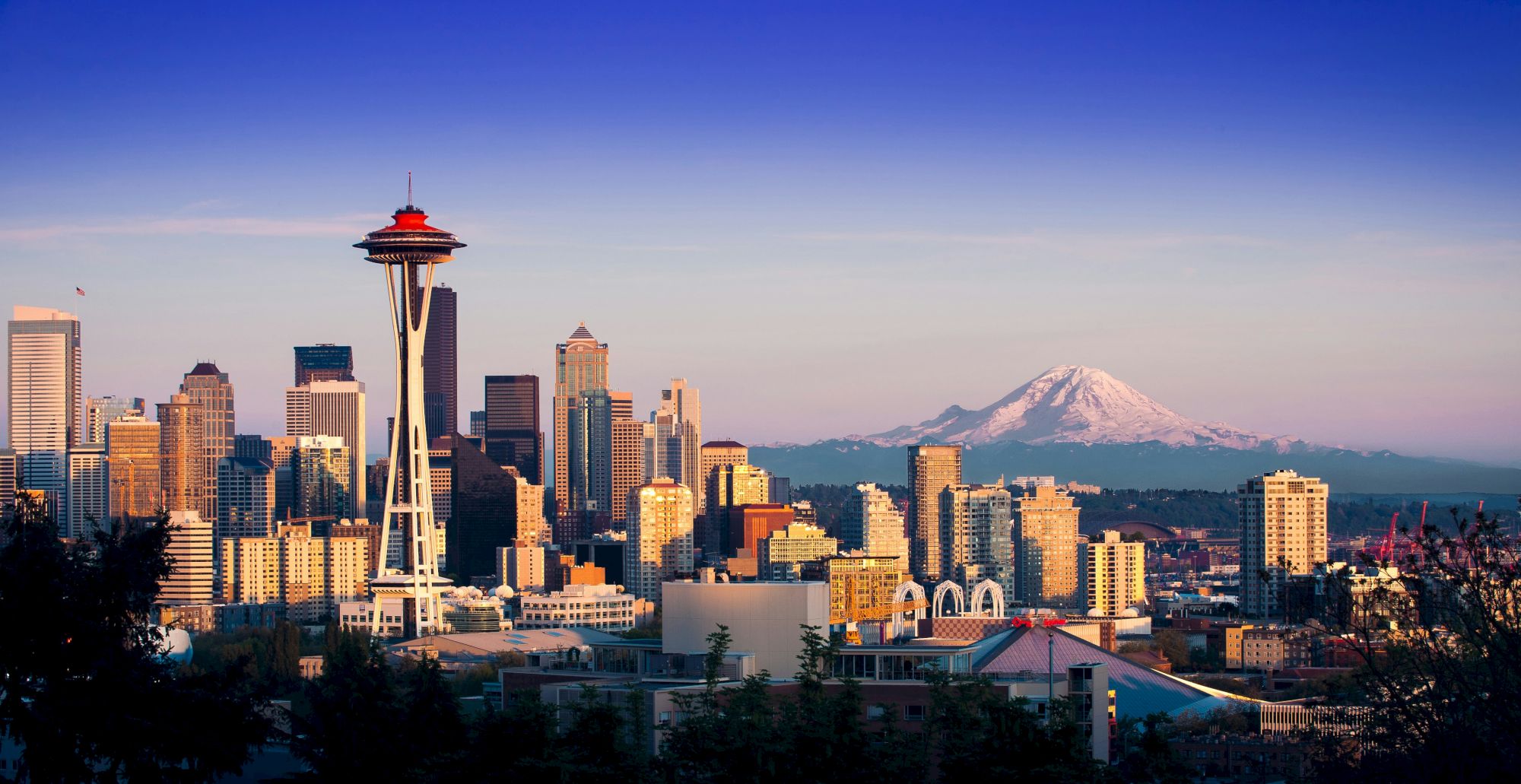 Seattle skyline with the Space Needle, tall buildings, and Mount Rainier in the distance; a clear blue sky above, city lights starting to glow.