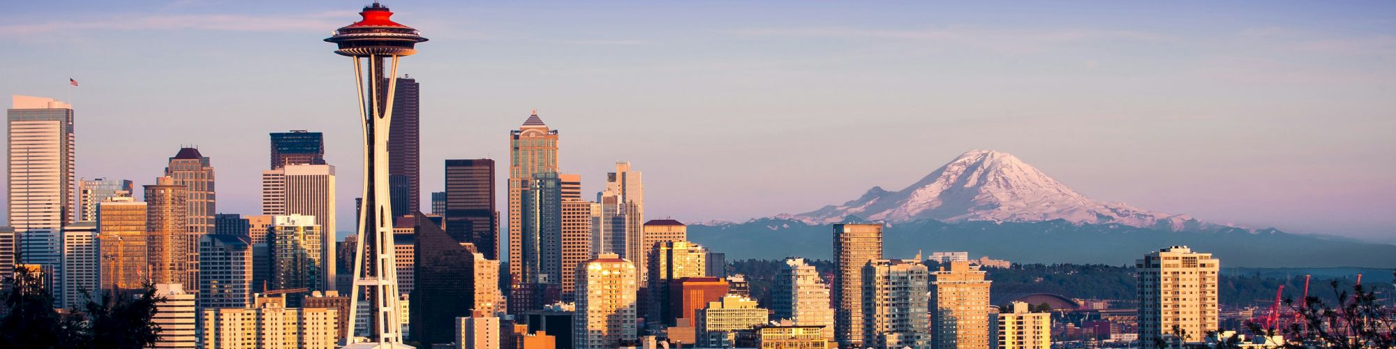 Seattle skyline with the Space Needle, tall buildings, and Mount Rainier in the distance; a clear blue sky above, city lights starting to glow.