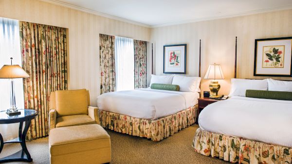 Two double beds in a hotel room with floral bed skirts, a chair and ottoman, two bedside lamps, floral curtains, and framed wall art.