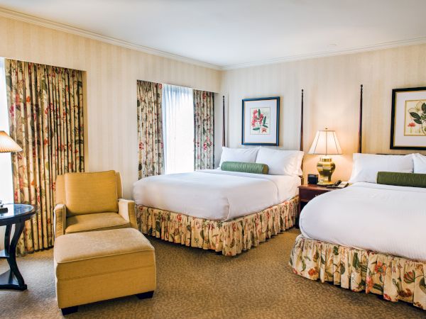 Two double beds in a hotel room with floral bed skirts, a chair and ottoman, two bedside lamps, floral curtains, and framed wall art.