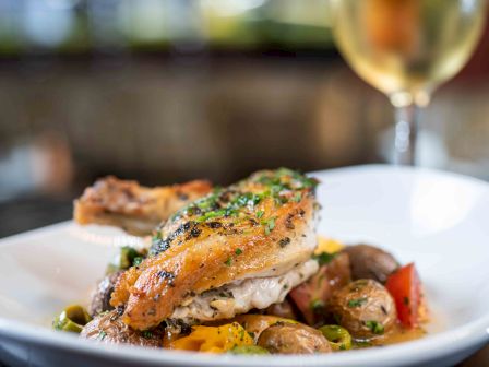 A plated dish featuring seared chicken on a bed of saut&eacute;ed vegetables (potatoes, peppers, peas) with herbs; wine glass in background.