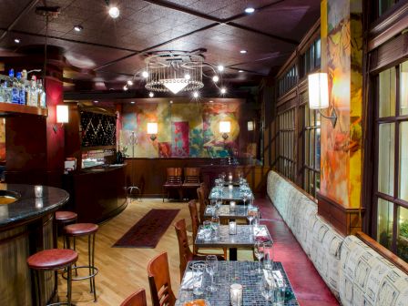 A stylish restaurant interior with a long dining table set for multiple guests, warm lighting, colorful walls, bar area, and tall windows.