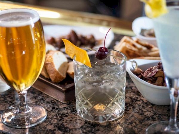 A bar setup with assorted drinks: a tall beer, a crystal old-fashioned glass with a cherry garnish, and a blue cocktail with a lemon twist, plus plates of appetizers in the background.