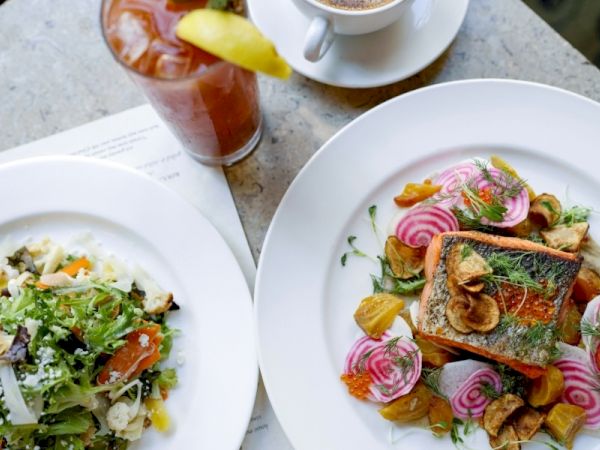 A pair of plates with food: left plate seems like a salad with greens and cheese, right plate features a fish fillet on chips with pickled onions and herbs, plus a glass of iced tea and a cup of coffee in the background.
