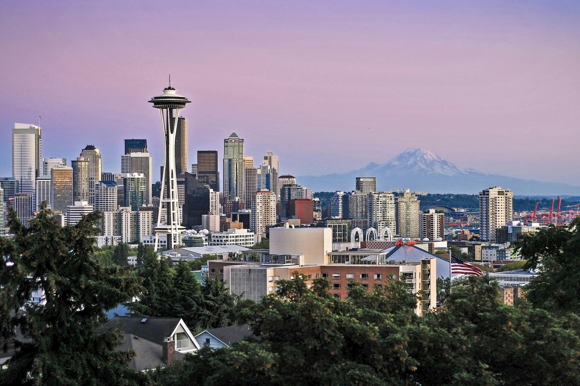 Seattle skyline with the Space Needle, modern buildings, trees in foreground, and distant mountains under a pinkish sky.