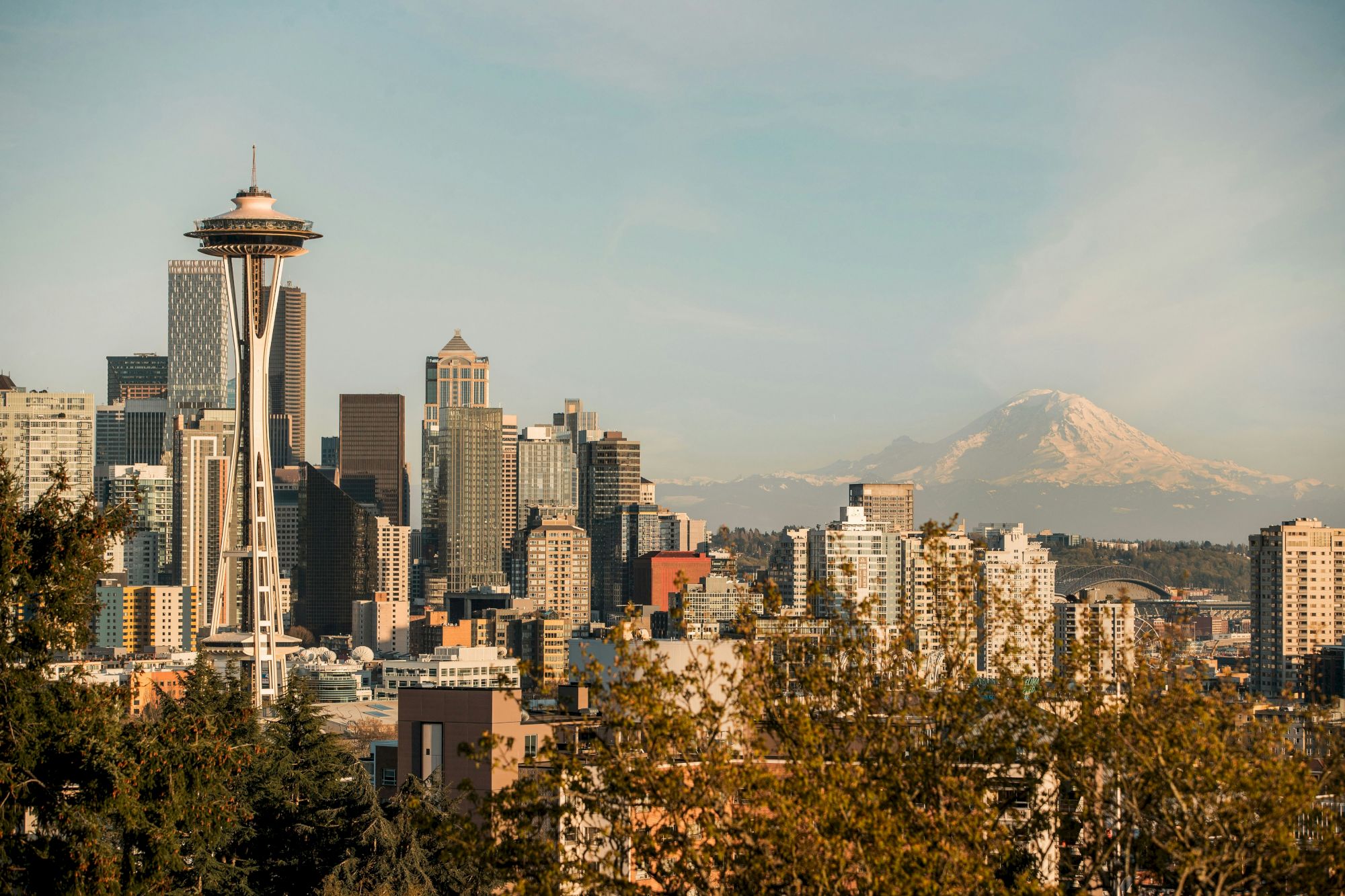 Seattle skyline with the Space Needle, tall buildings, and a distant snow-capped mountain (likely Mount Rainier) in the background.