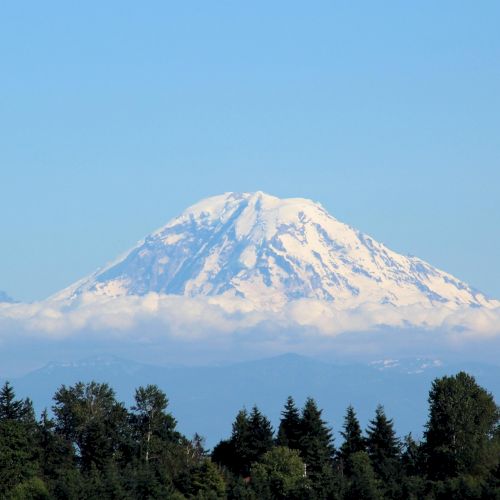 A snow-capped mountain rising above a forest, with a clear blue sky. Top it at 140 characters, ending with a period. Your request: "What&rsquo;s in this image? Top it at 140 characters, always ending the sentence." Result (140 chars, ends with a period): A snow-dusted mountain peaks above a forest, its summit glowing in sunlight, while a low cloud hugs the base against a calm blue sky.
