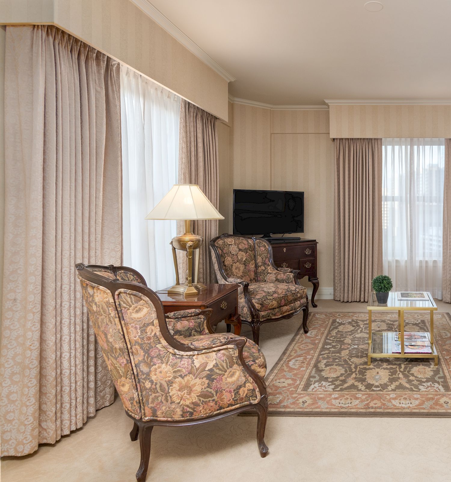 A comfortable living room with cream walls, beige sofa, patterned armchairs, fireplace screen, lamps, and a glass coffee table&mdash;bright curtains frame the windows.