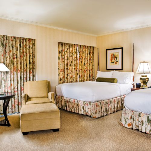 A hotel room with two queen beds, floral curtains, a small seating area, and soft lighting&mdash;cozy, coordinated decor.