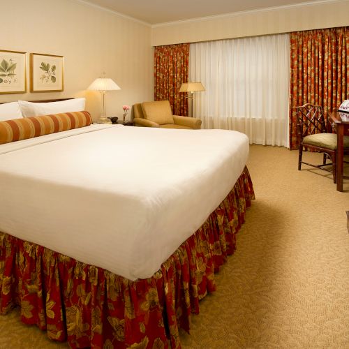 A spacious hotel room with a large bed, floral bed skirt and pillows, framed botanical art, a seating area, desk, lamps, and a TV, with red patterned curtains.