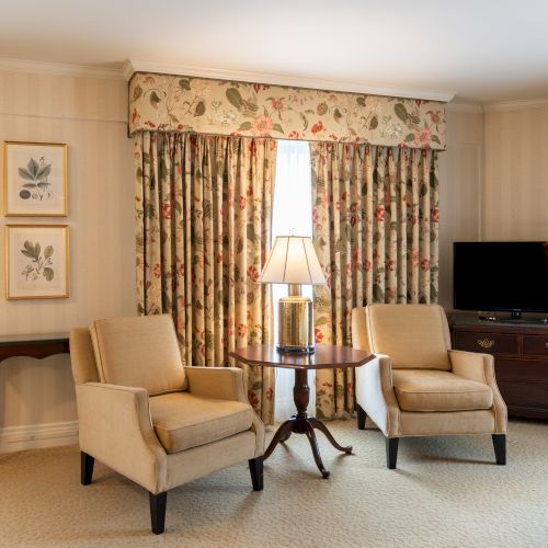 Cozy hotel suite with floral drapes, two beige armchairs around a small table, a lamp, side cabinet, and a TV on a dresser. End.