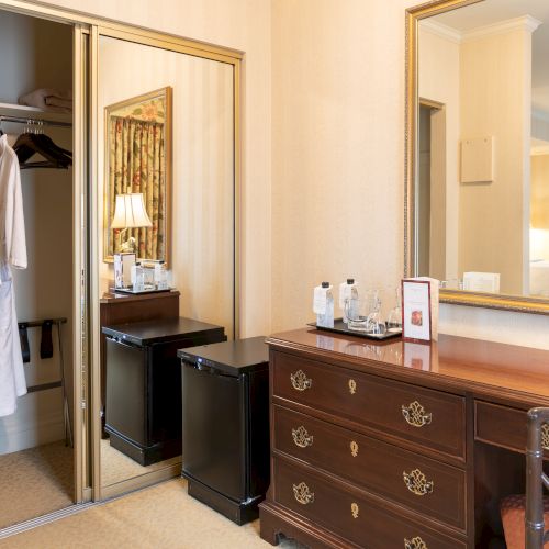 A hotel room closet with robes, a dresser, a mirror, a lamp, and a small safe or minibar area; tidy and classic, hotel style.