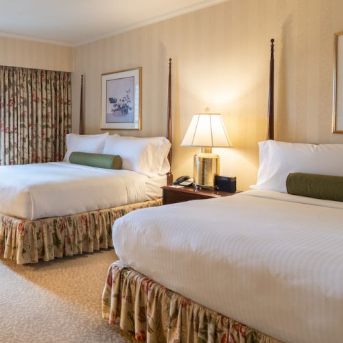 A hotel room with two queen beds, floral bed skirts, beige walls, a seating chair by the window, and a nightstand with lamp and framed art. End.