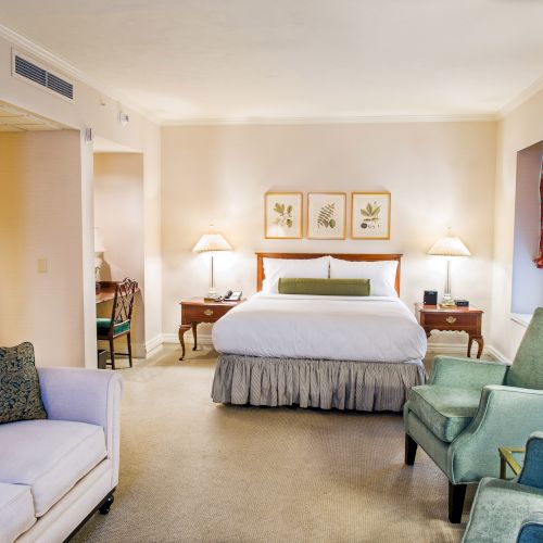 A spacious hotel-style bedroom with a four-poster bed, two nightstands, a seating area with a sofa and green chairs, and warm lighting. End.