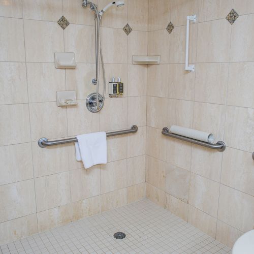 A wheelchair-accessible bathroom with tiled walls, multiple grab bars, a showerhead, built-in shelves, a toilet, and a small drain on the floor, all finished in light beige.