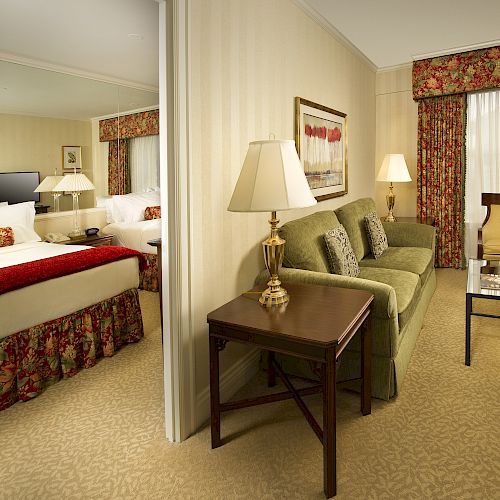 A hotel suite living area with a sofa, coffee table, lamps, and a view into a bedroom with two beds and floral window treatments.