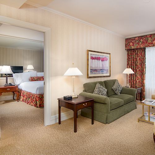 A hotel suite living area with a green sofa, side tables, lamps, a coffee table, and a flat-screen TV; connected to a bedroom with a bed.