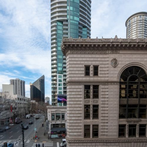 A city street scene with tall modern buildings and a historic stone facade in the foreground, blue sky and scattered clouds above.