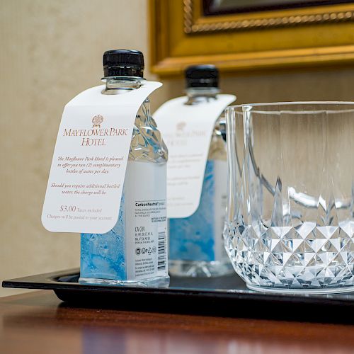 A hotel-supplied glass pitcher and crystal tumbler set on a silver tray, with bottled water and two upside-down glasses on a wooden table.